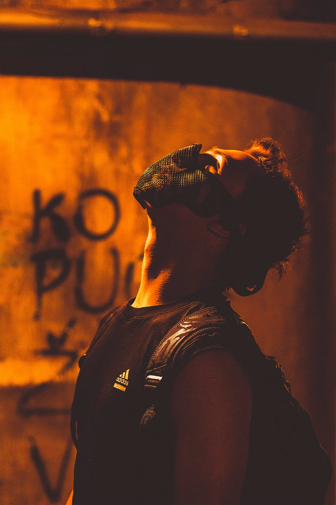 Person in a black athletic shirt with a backpack, wearing a mesh mask, standing in a dimly lit area with orange lighting and graffiti on the wall.