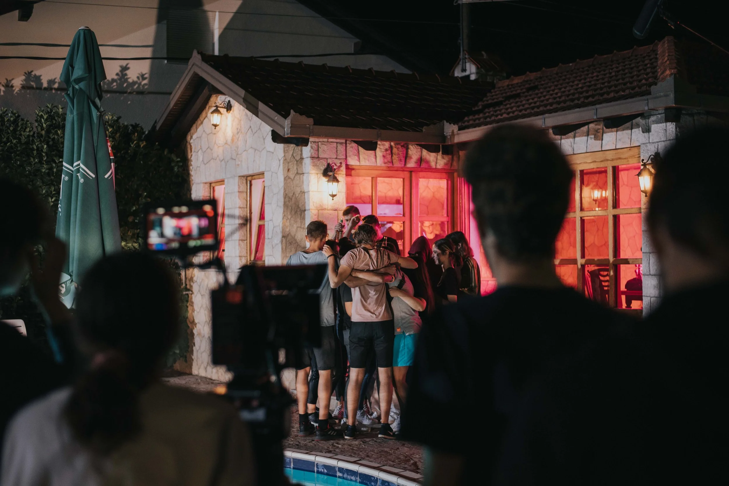 People gather outside a house with pink and red lighting, some hugging, at night, with a camera recording.