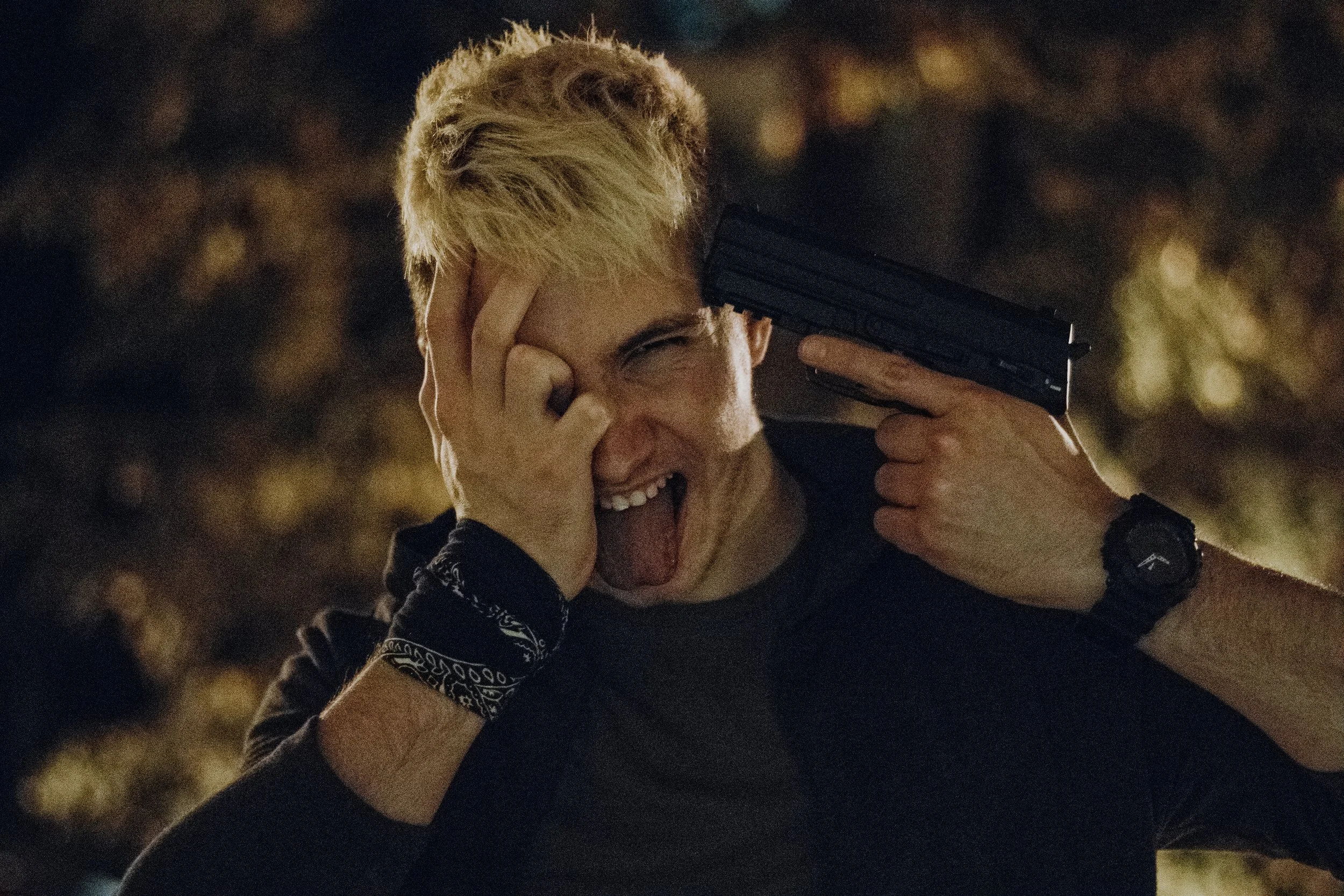A young man with blonde hair making a playful expression, holding a gun to his head with his left hand and covering his right eye with his right hand, sticking out his tongue. He is wearing a black shirt, a watch on his left wrist, and a black bandan