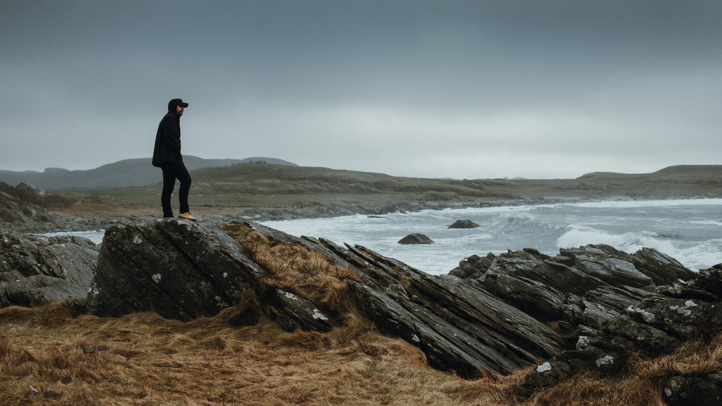 Person standing on large rocks by the ocean on a cloudy day with hills in the background.