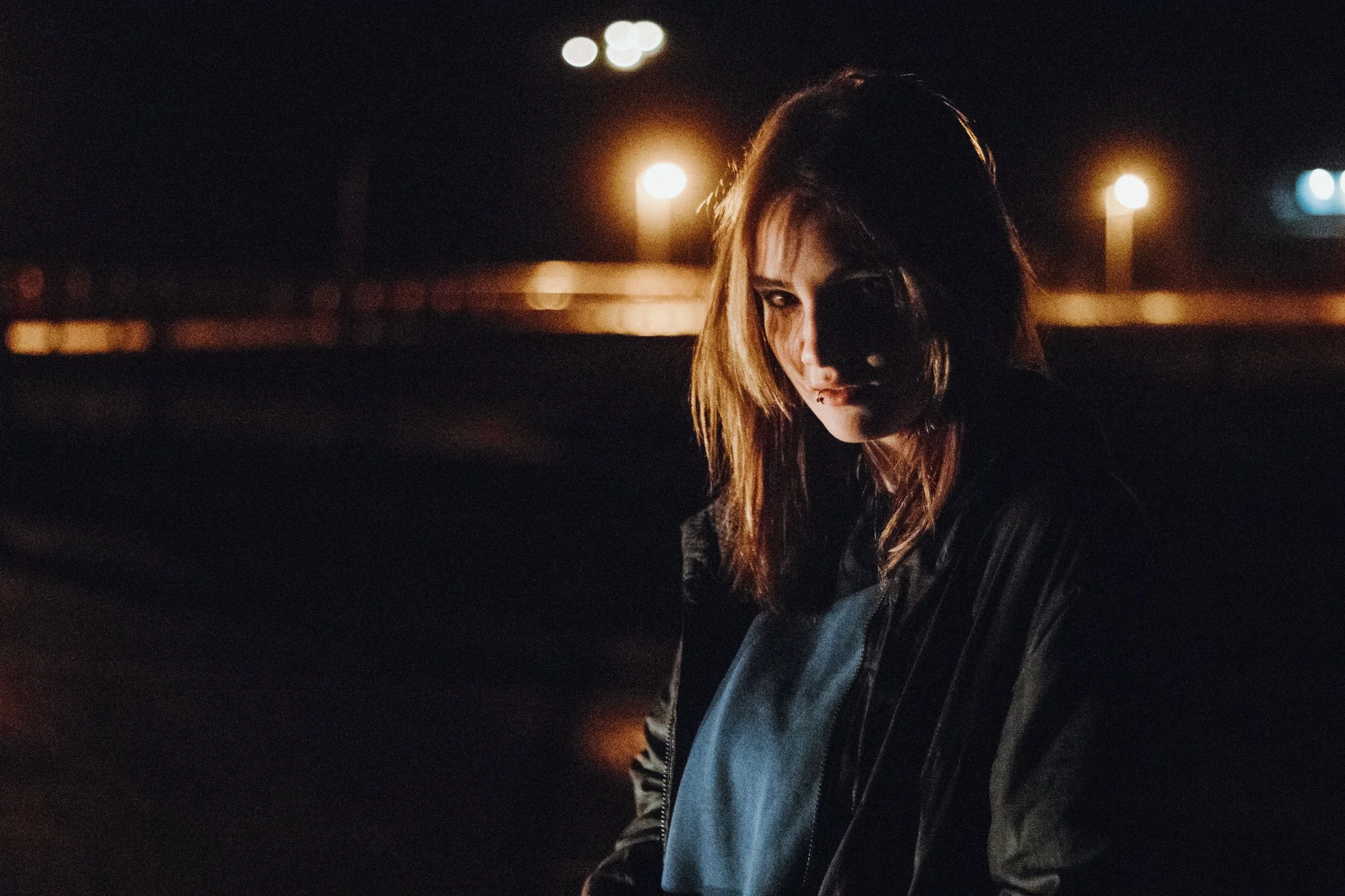 A woman with brown hair in a dark outfit sitting outdoors at night, illuminated by a few streetlights in the background.