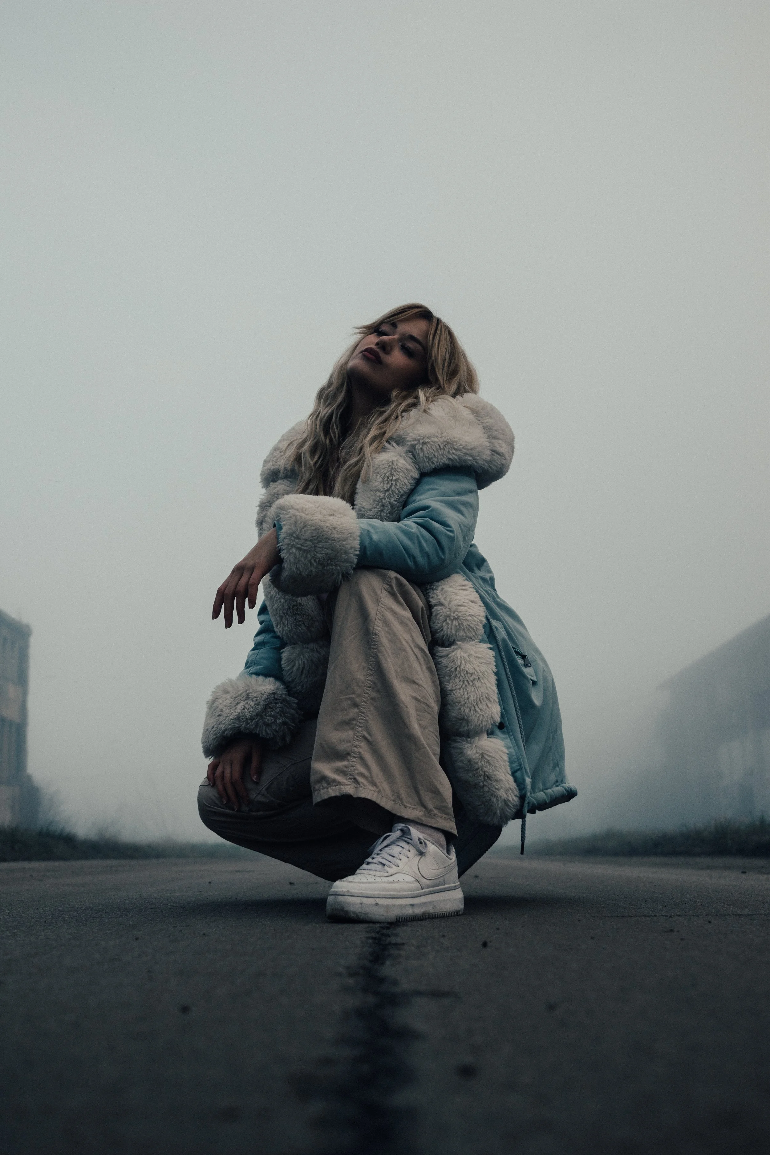A young woman crouching on an empty road in a foggy environment, wearing a blue winter coat with fur trim, tan pants, and white sneakers.