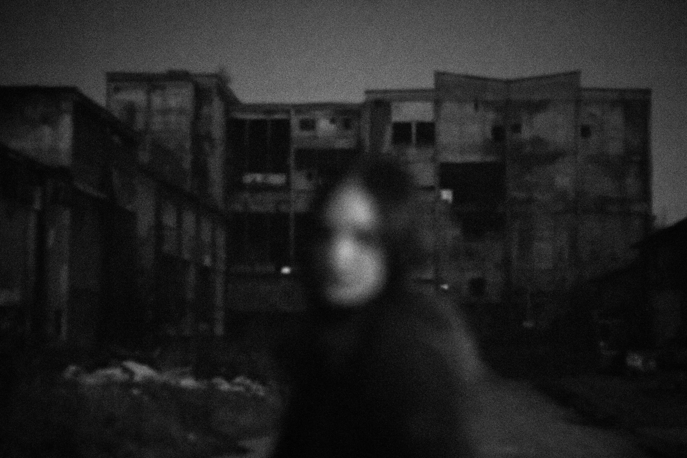 A blurry black and white photo of a person in the foreground with a large, multi-story building in the background at night.