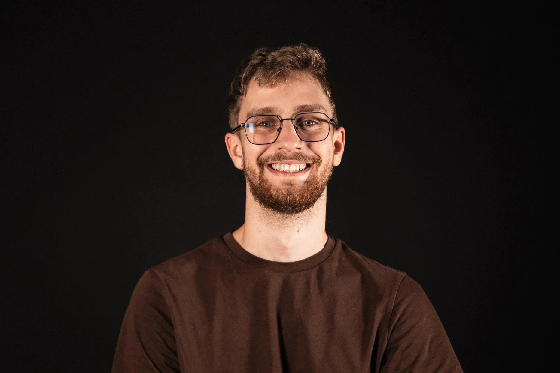 A young man with glasses, a beard, and short brown hair smiling against a black background.