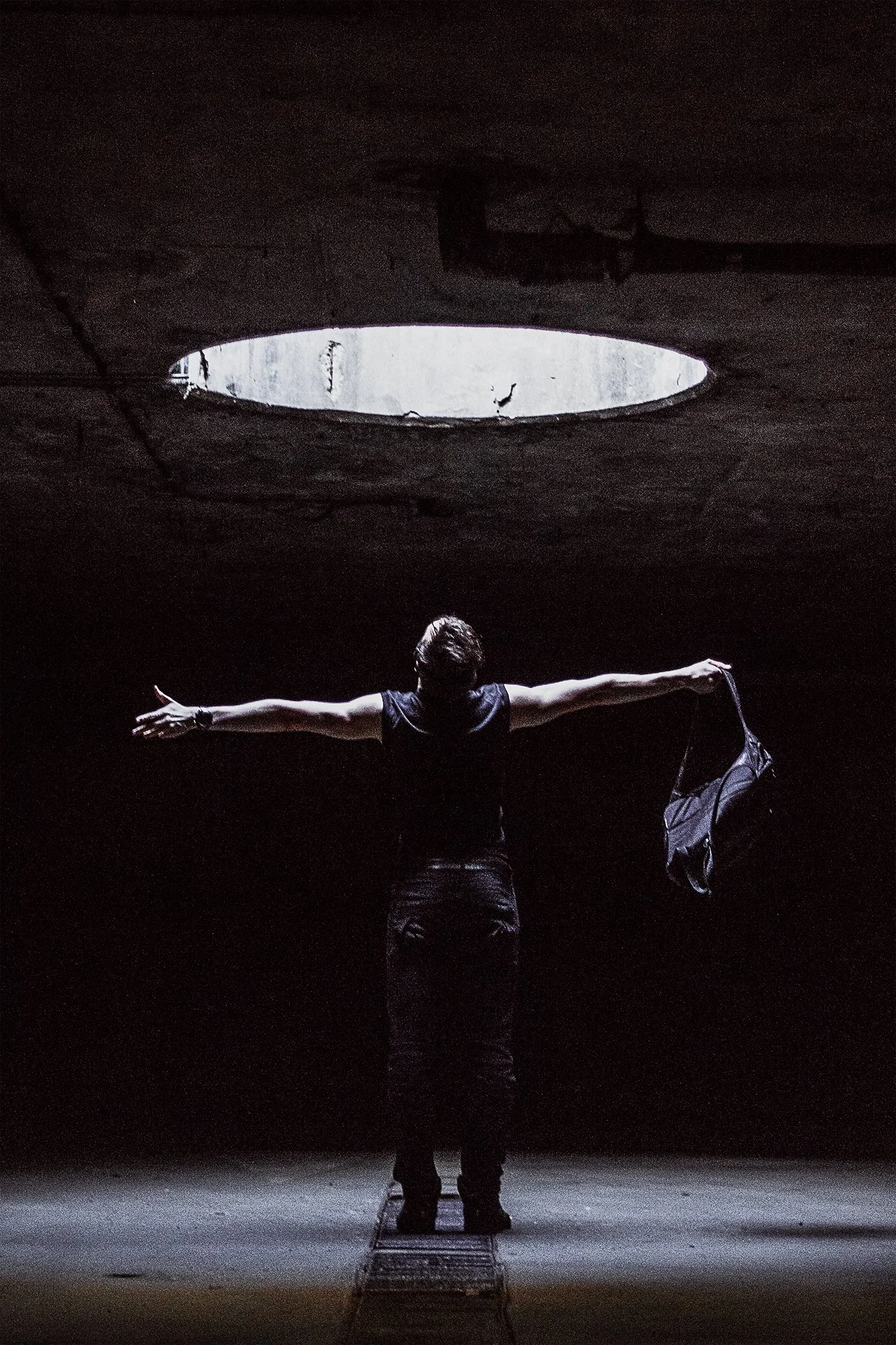 Person standing with arms outstretched in a dark, empty space with a circular opening in the ceiling above.