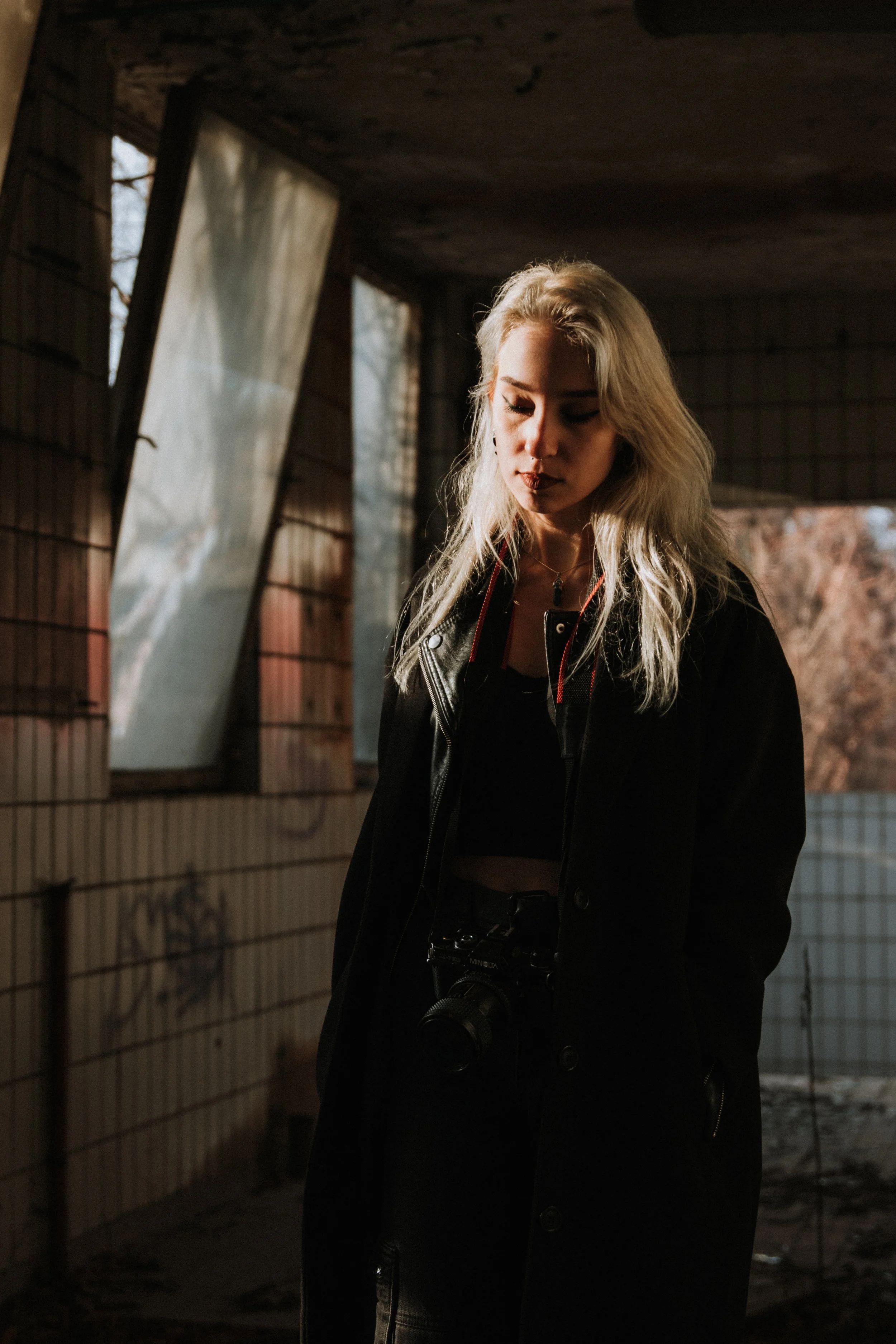 A young woman with long blonde hair standing indoors near a window with broken or open panels, wearing a black coat and a camera around her neck, looking down.