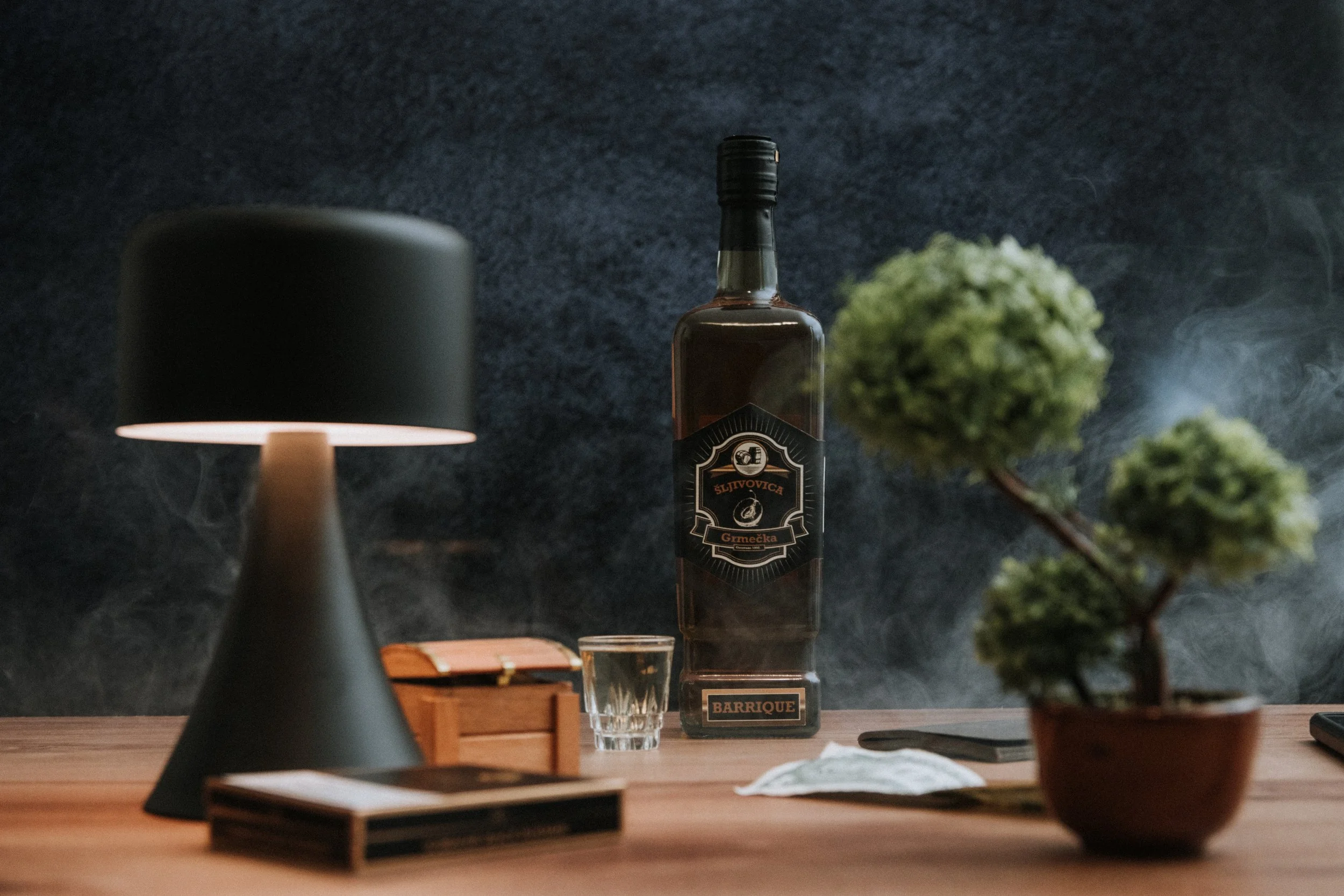 A bottle of liquor, a glass, a small potted plant, a wooden box, a book, and a table lamp on a wooden table against a dark background.