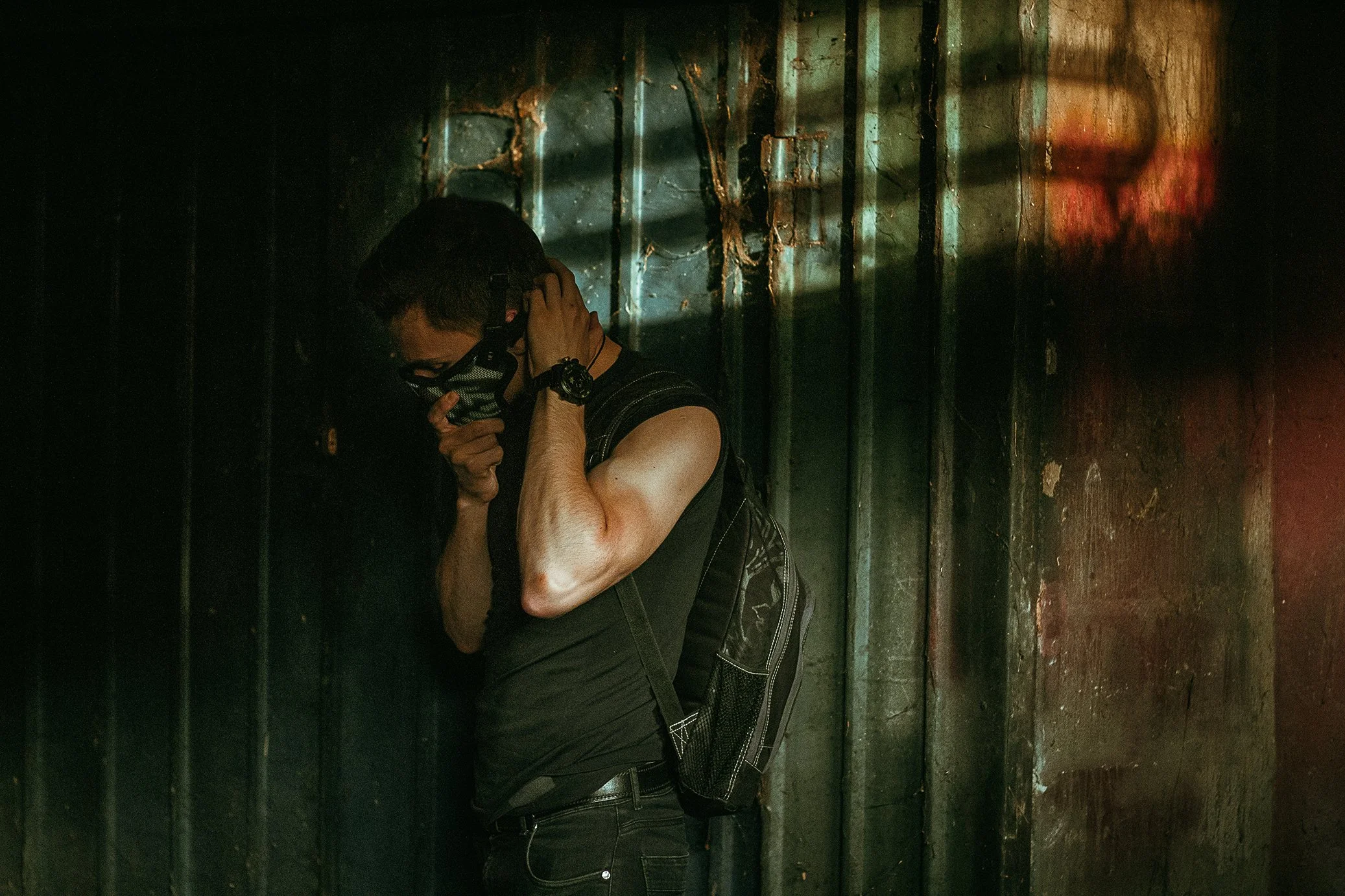 A person wearing a face mask, black sleeveless shirt, and carrying a black backpack, leaning against a dark, weathered wall, with one hand on their face and the other touching their neck, surrounded by shadows and dim lighting.