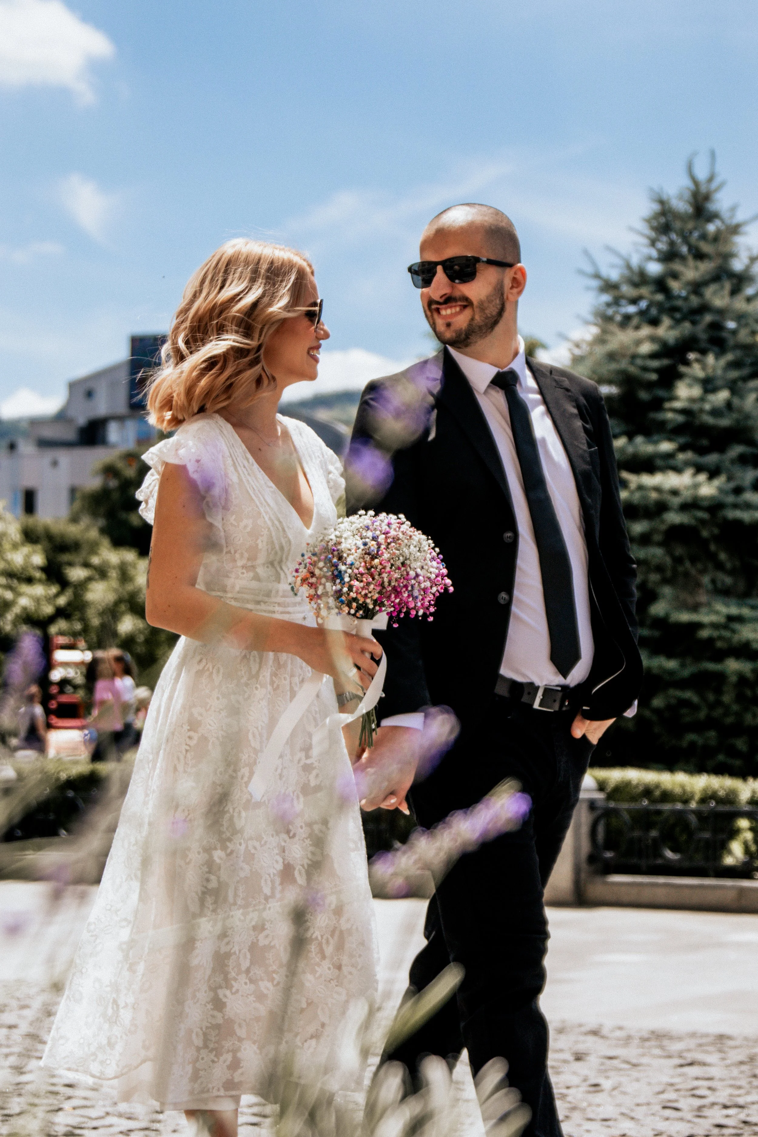 A smiling bride and groom walking outdoors on a sunny day, holding hands. The bride is wearing a lace white dress and sunglasses, holding a bouquet of pink and white flowers. The groom is wearing a black suit, white shirt, black tie, and sunglasses. 