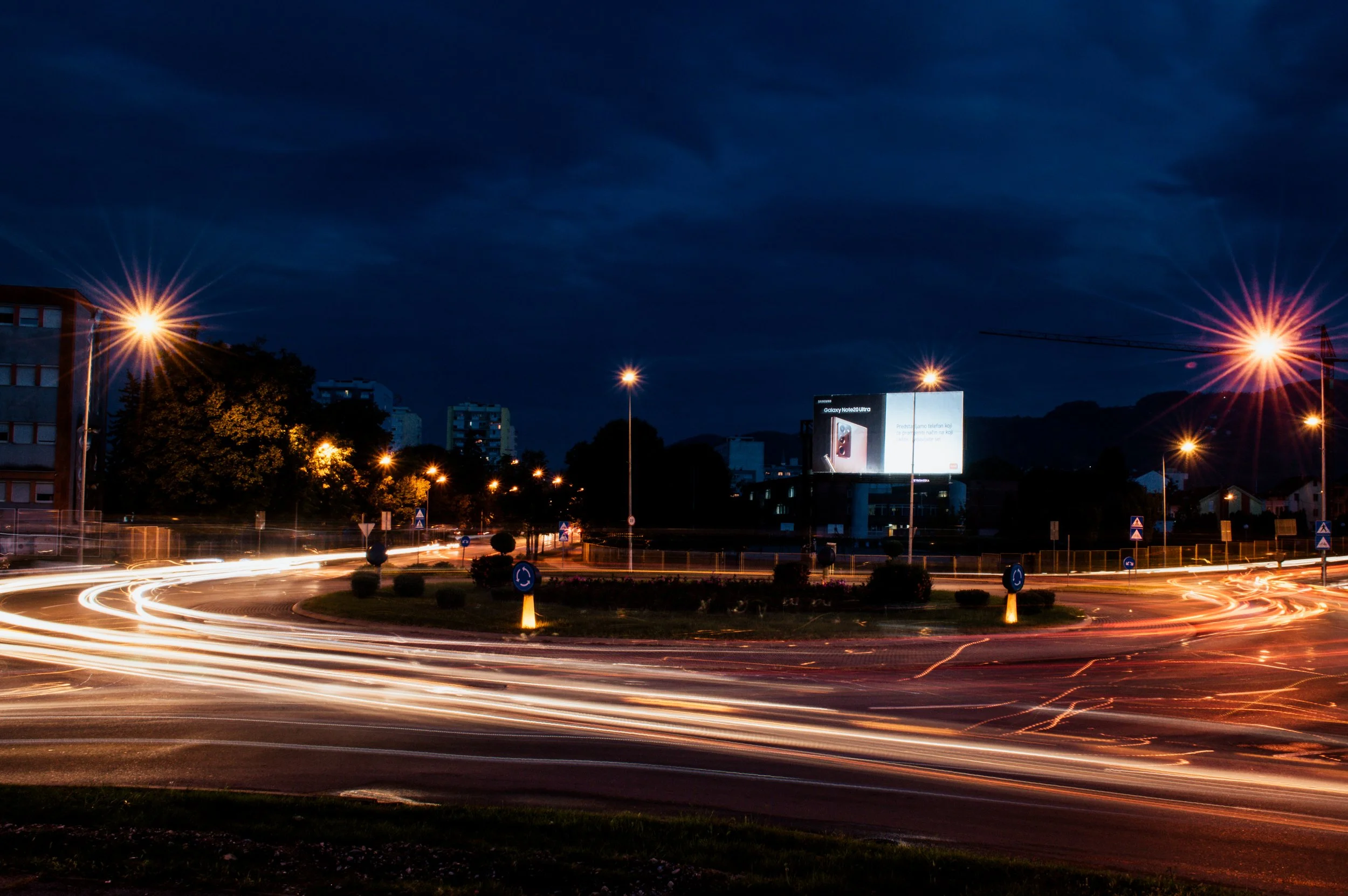 Nighttime city street roundabout with illuminated light trails from passing vehicles and streetlights, buildings, and a billboard in the background.