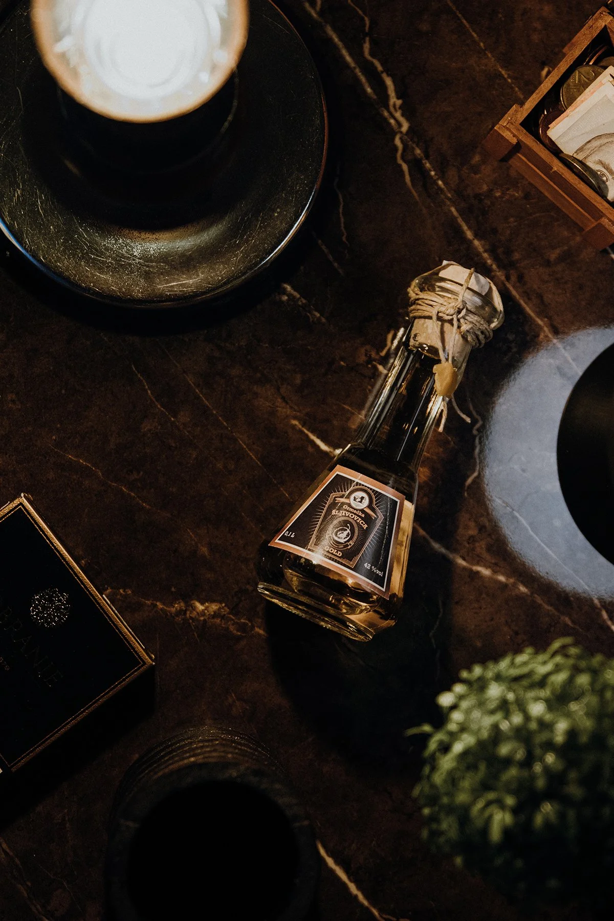 A bottle of liquor with a cork, placed on a dark marble countertop, surrounded by small boxes, a brass tray with a cup, and a small potted green plant.