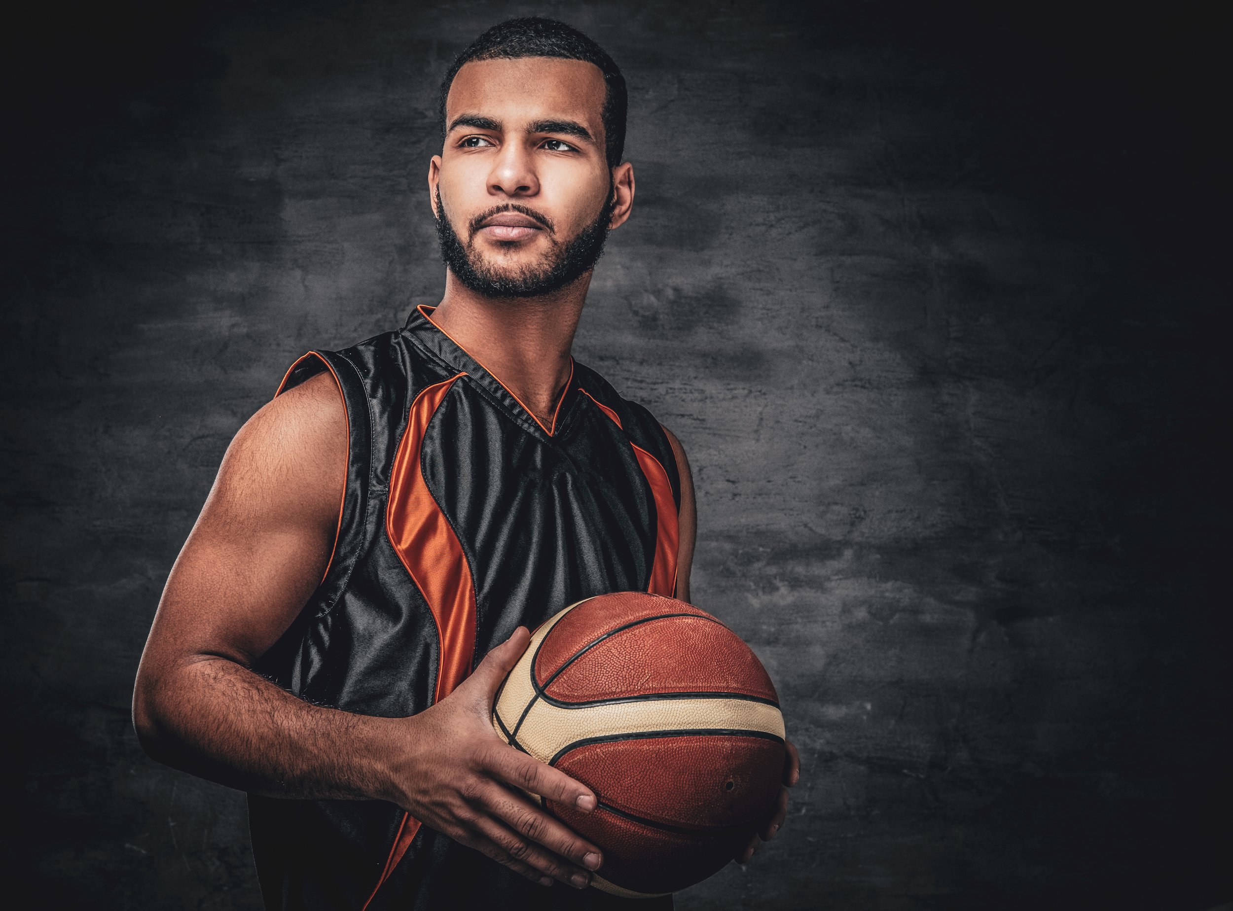 A young man with a beard and short hair holds a basketball, wearing a black and orange basketball jersey, looking off to the side against a dark textured background.
