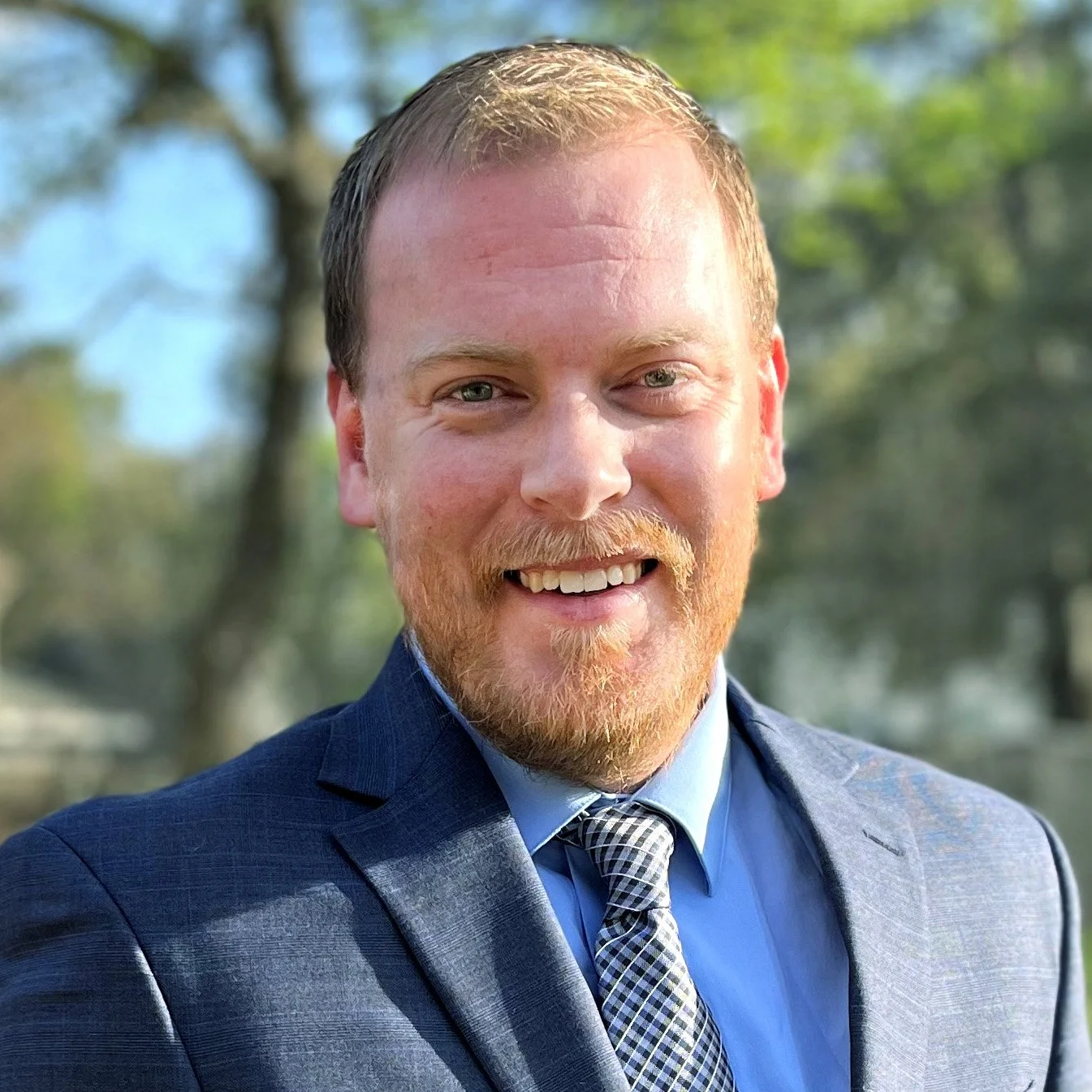 A man with light skin, short reddish hair, and a beard, smiling outdoors with trees in the background, wearing a blue suit, blue shirt, and a patterned tie.