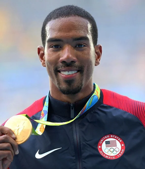 Athletic man holding gold medal, wearing USA Olympic team jacket with Nike logo, smiling with short hair and goatee, colorful Olympic medal ribbon around neck.