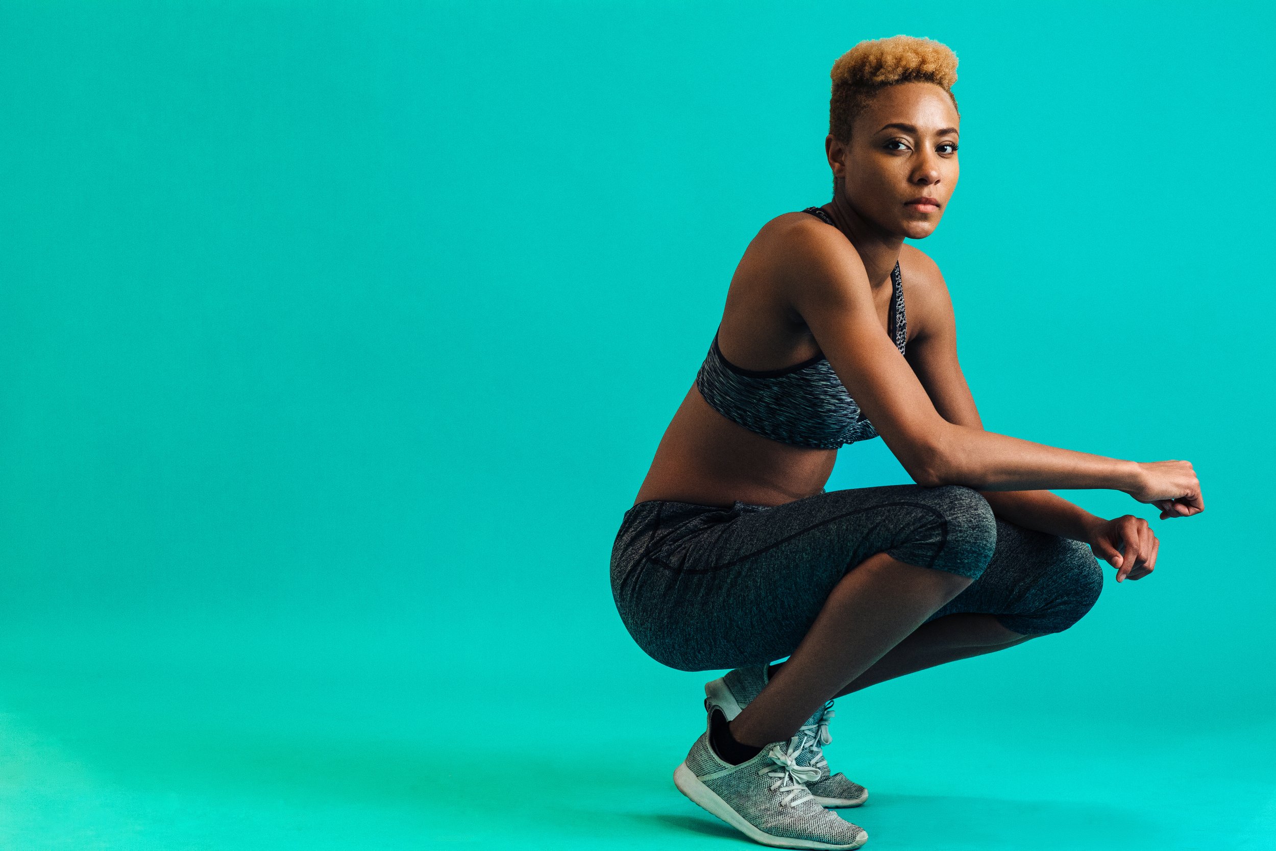 A woman with short blonde hair squat down on a teal background, wearing a black and gray sports bra, dark gray athletic leggings, and gray athletic shoes.