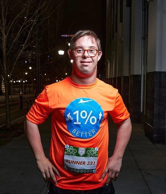 A man with glasses and light brown hair, wearing an orange running shirt with a blue circular logo, standing outdoors at night. He has his hands on his hips and a race bib with the number 321.