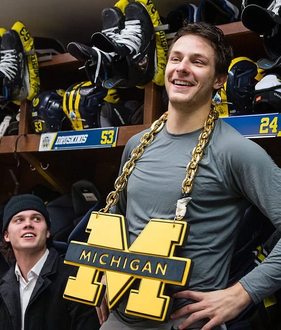A young man smiling and wearing a large gold chain with a Michigan logo pendant in a hockey locker room, surrounded by hockey equipment and jerseys.