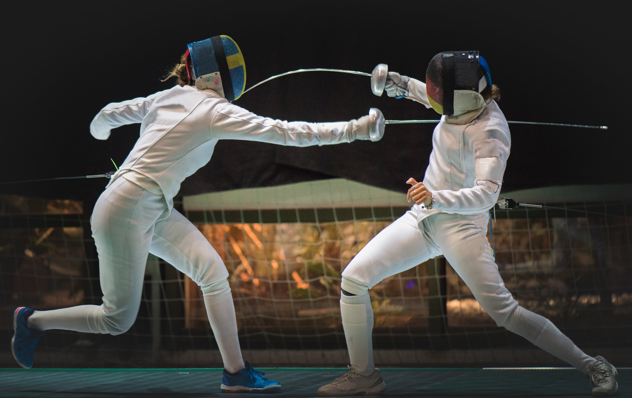Two female fencers sparring on a fencing strip at night, wearing protective gear and masks, engaged in a foil match.