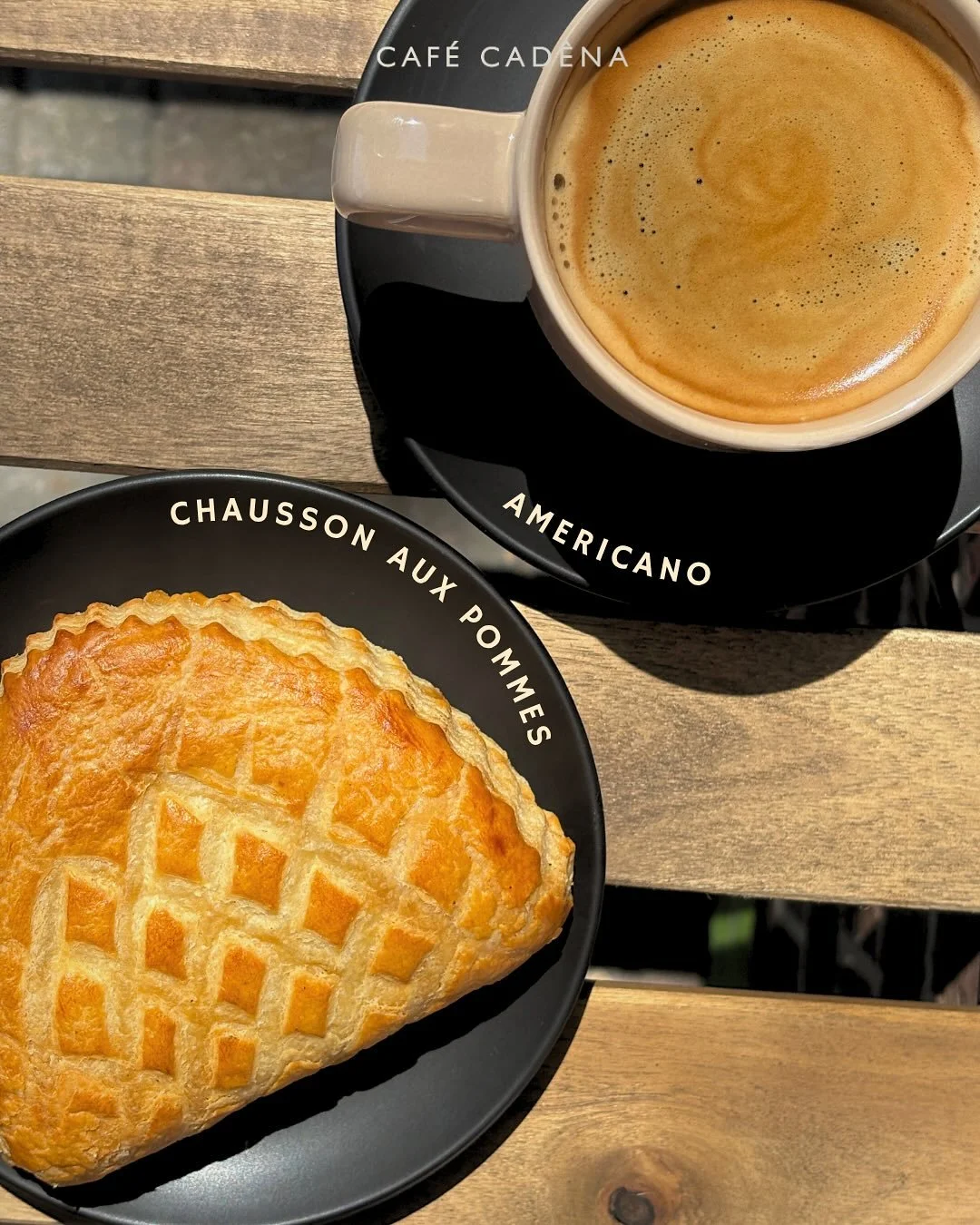 Une tasse de café américain avec une crème mousseuse et un chausson aux pommes doré sur une assiette noire avec des inscriptions en français et en anglais, posés sur une table en bois