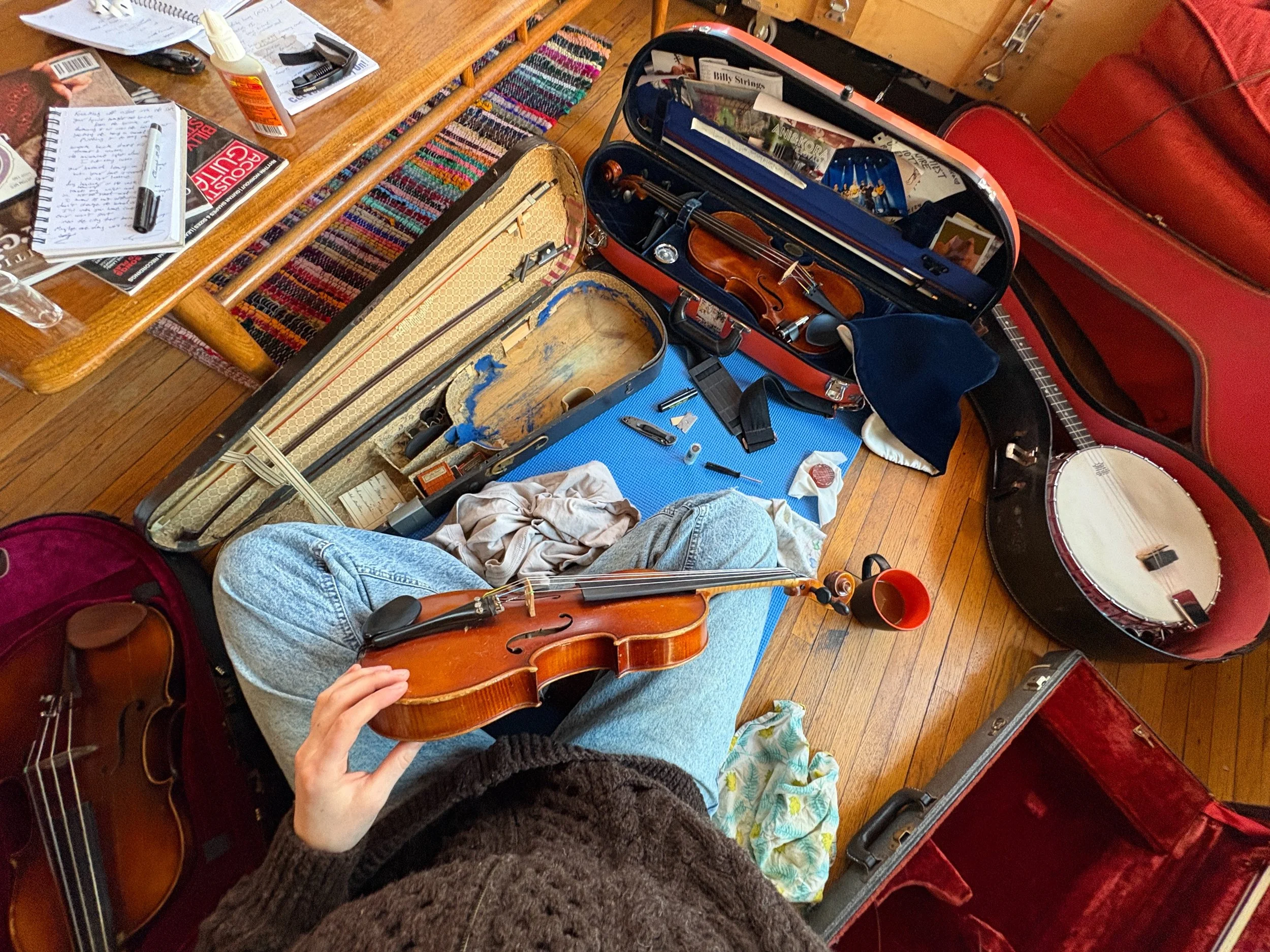 person holding a violin while sitting on a blue mat, casea and music books open nearby on a wooden floor.
