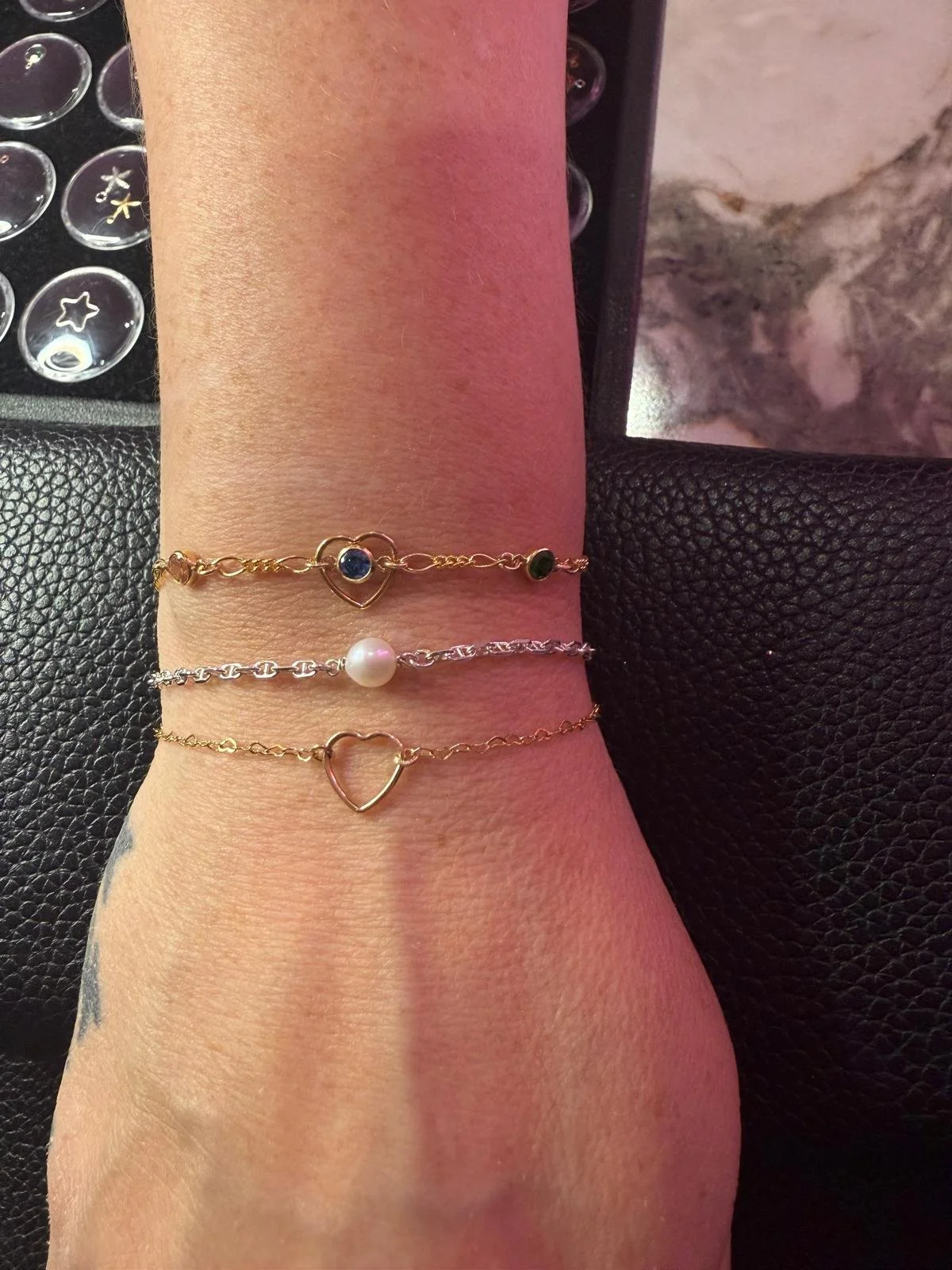Three delicate gold and silver permanent welded bracelets with charms on a person's wrist, including heart shapes, a pearl, and birthstones.