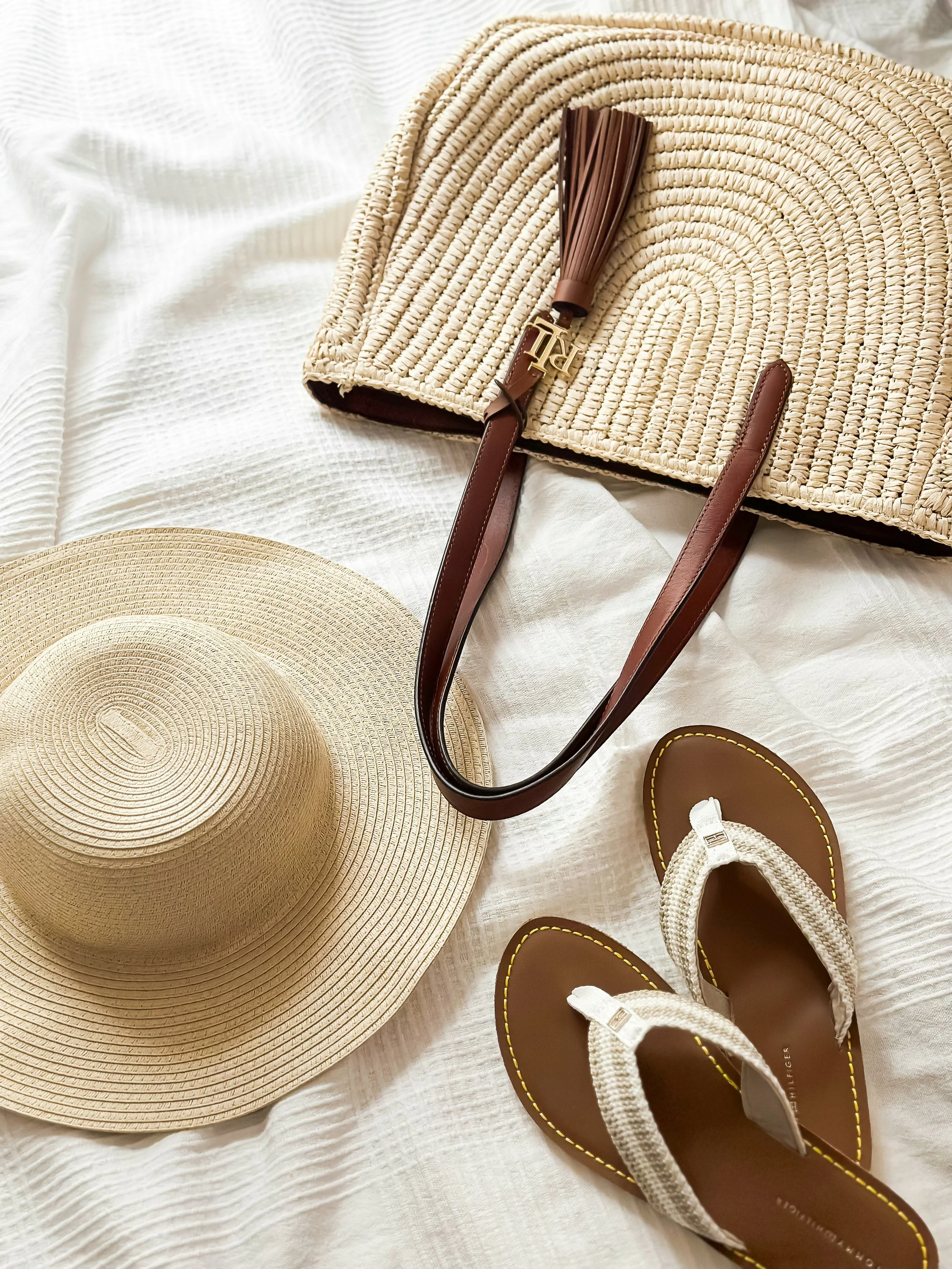 A wide-brimmed straw sun hat, a pair of brown and white sandals, a patterned woven tote bag with leather handles and a tassel, all arranged on a white bedspread.