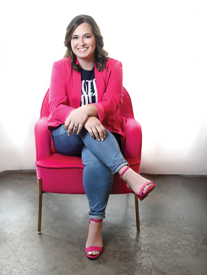Amanda sits in the Pink Chair wearing a teeshirt that reads "Foster Confidence" under a pink blazer, with jeans and pink shoes