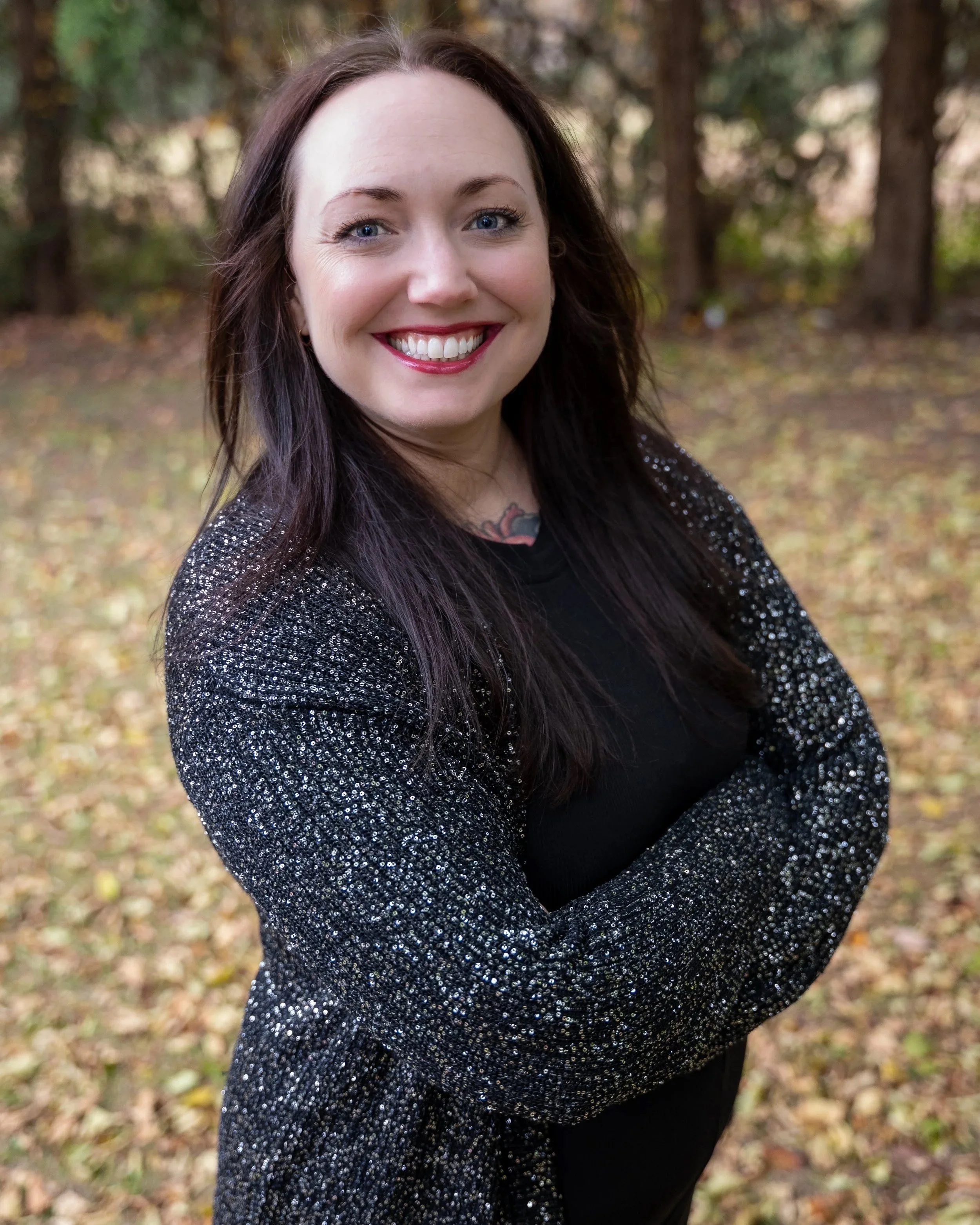 A woman with long dark hair, wearing a sparkly black jacket and black shirt, smiles outdoors in a wooded area with fallen leaves.