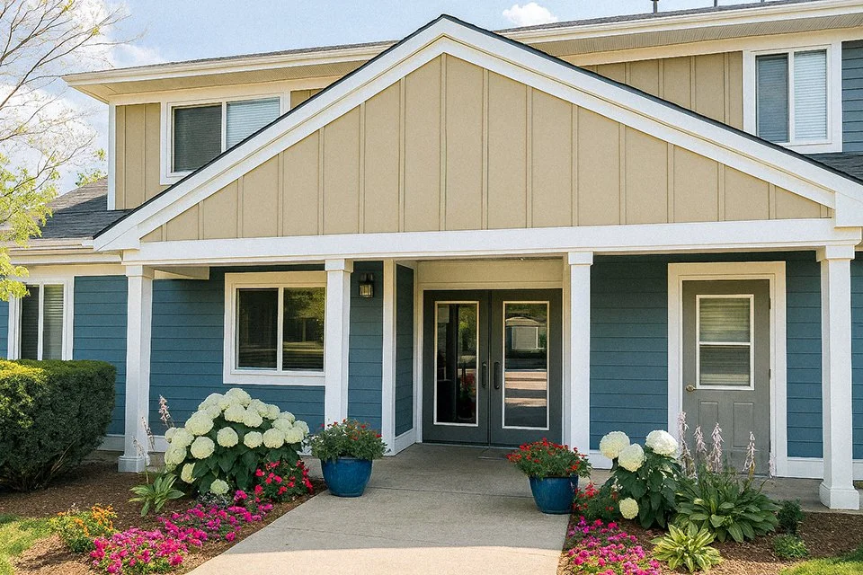 Front view of a two-story house with blue siding and beige gable, white trim, covered porch with white columns, glass double doors, and flower pots with white and pink flowers outside.