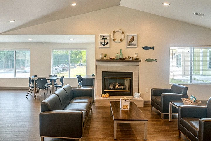 Living room with a fireplace, dark leather sofa and chairs, wooden coffee table, nautical decorations above fireplace, large windows, and a dining area in the background.