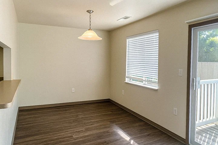 Empty dining area with hardwood flooring, a window with white blinds, sliding glass door leading to a balcony, and a hanging light fixture.