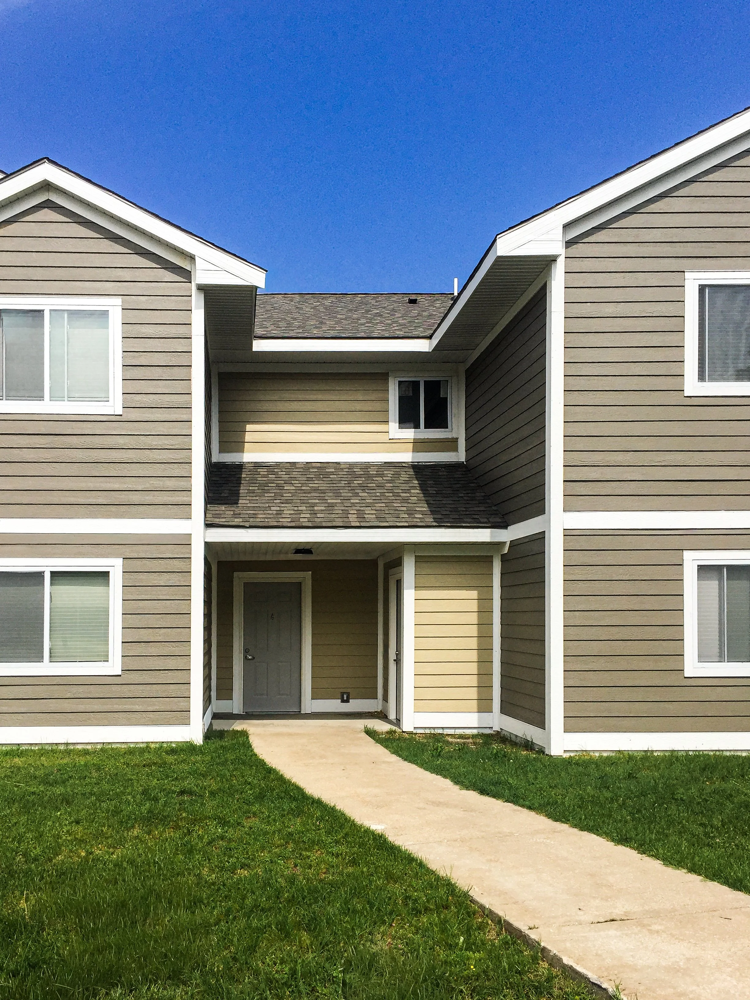 Front view of a multi-unit beige and brown apartment building with a concrete walkway leading to the main door, small grass lawn, white trim, and a clear blue sky.