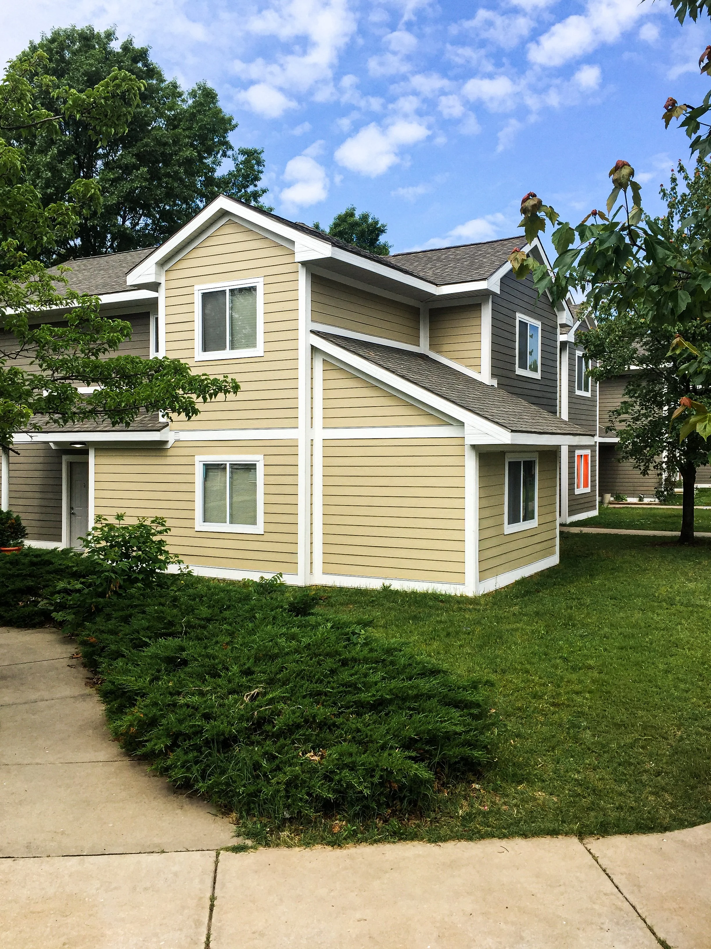 A two-story residential building with beige and gray exterior, white trim, multiple windows, surrounded by a green lawn, bushes, and trees, under a blue sky with scattered clouds.