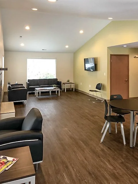 Spacious living room with black sofas, a wall-mounted TV, a small round dining table with black chairs, and a large window providing natural light.