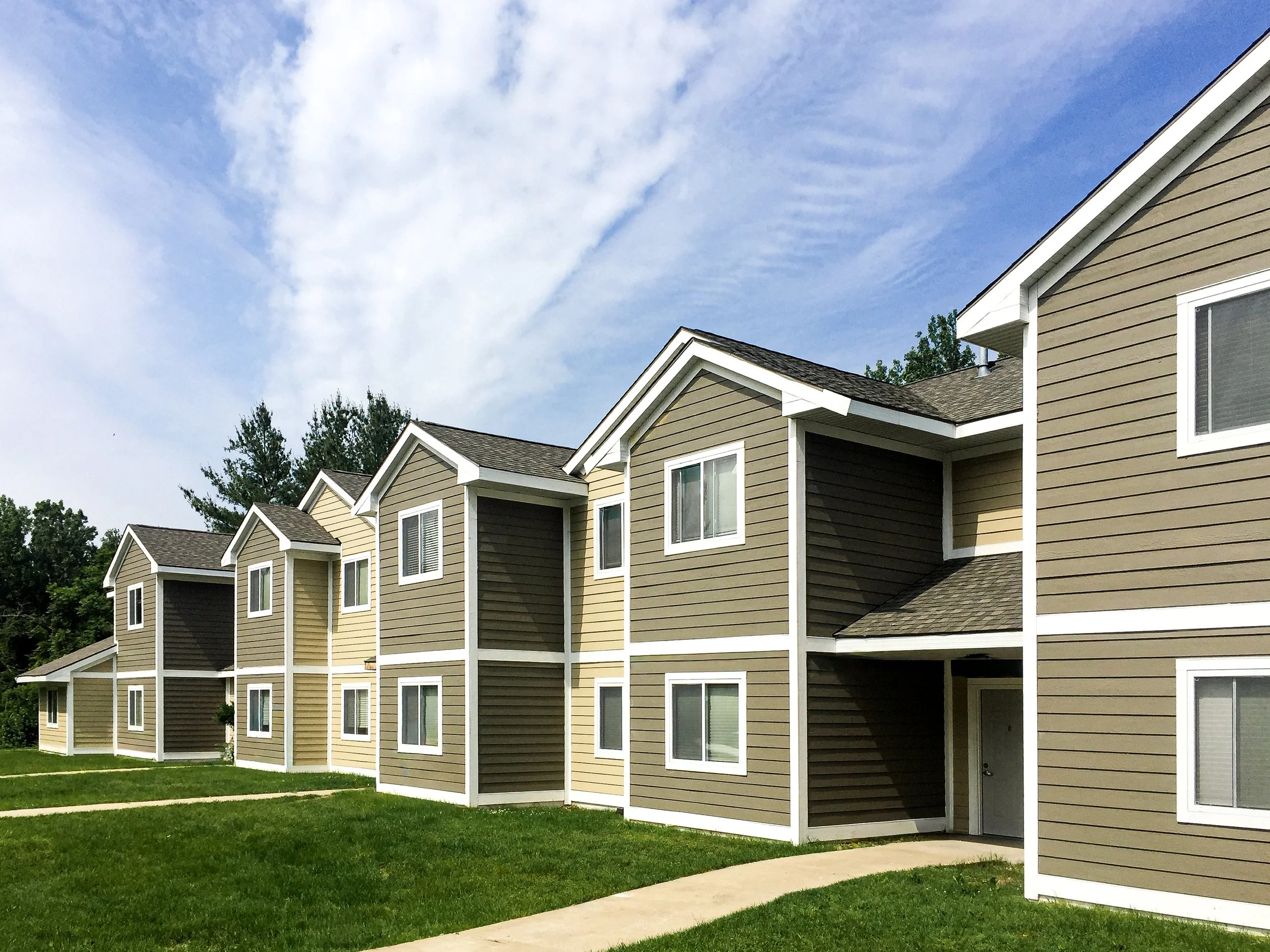 Multiple beige and brown apartment buildings with white trim, set against a partly cloudy blue sky and surrounded by green grass.
