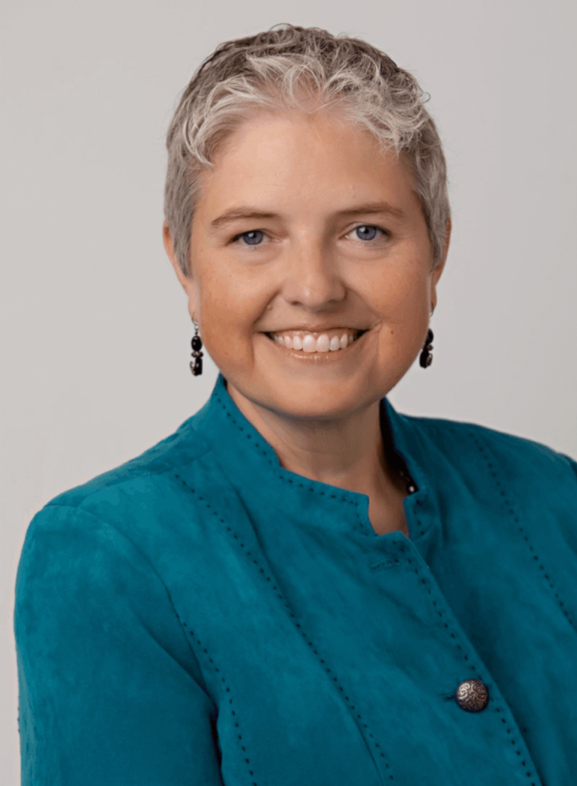 A middle-aged woman with short gray hair, smiling, wearing a teal jacket and black earrings, against a plain gray background.