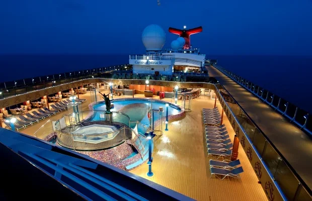 View of the deck on a cruise ship at night, featuring a pool, hot tubs, lounge chairs, and the ship's funnel in the background.