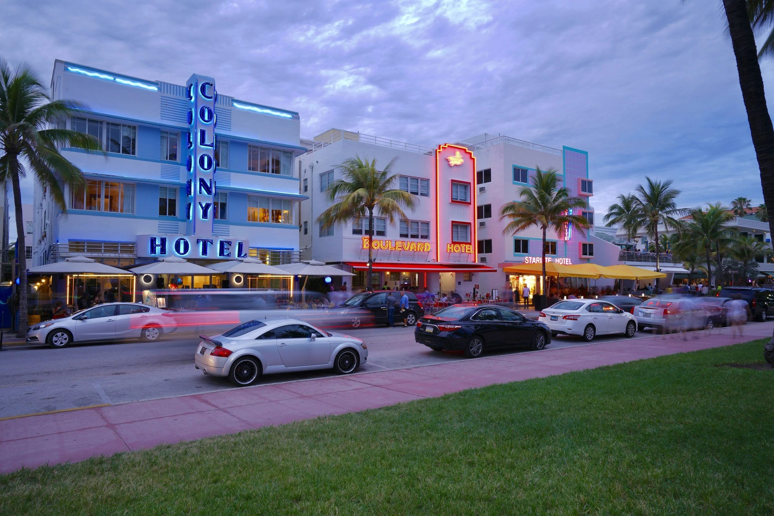 Night scene of a street with three colorful, neon-lit hotels with palm trees in front and cars parked along the curb.