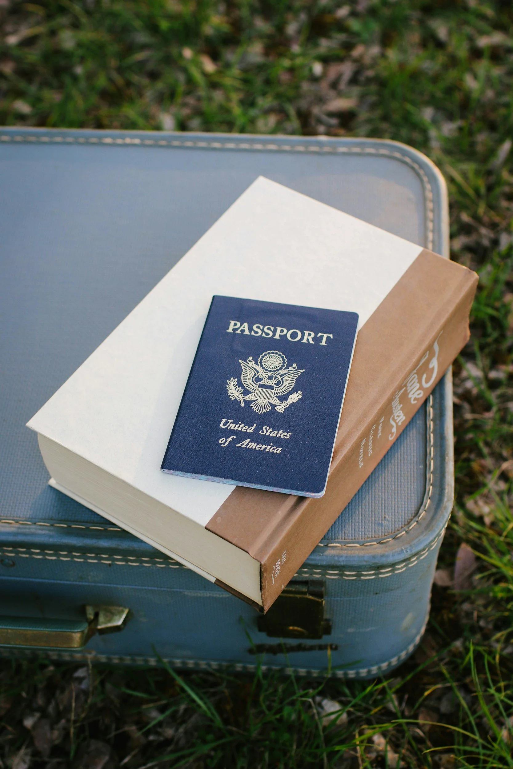 A United States passport on top of a closed cardboard box, resting on a light blue suitcase outside on grass.