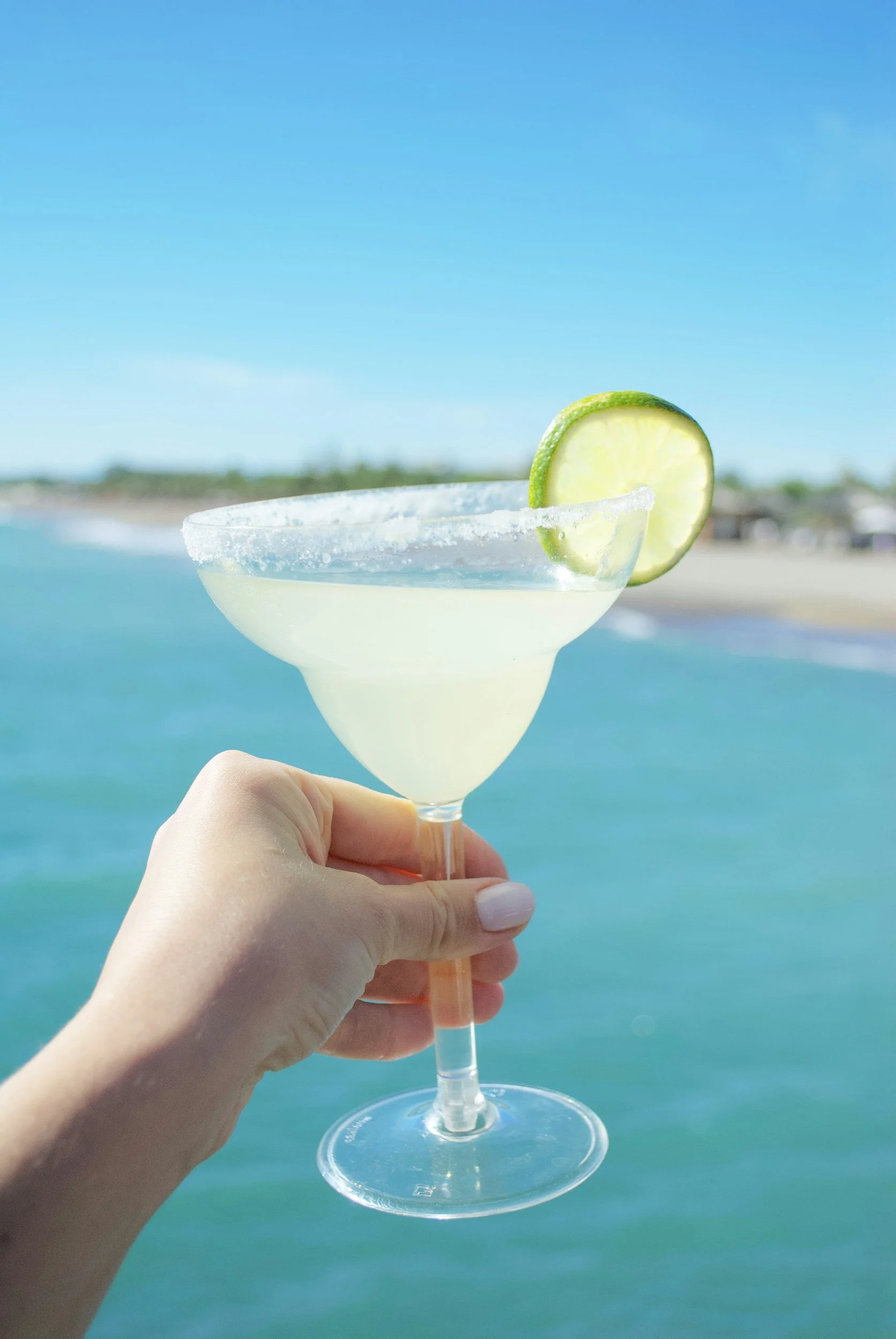 A hand holding a margarita glass with a salted rim and a lime wedge garnishment, with a beach and ocean in the background.