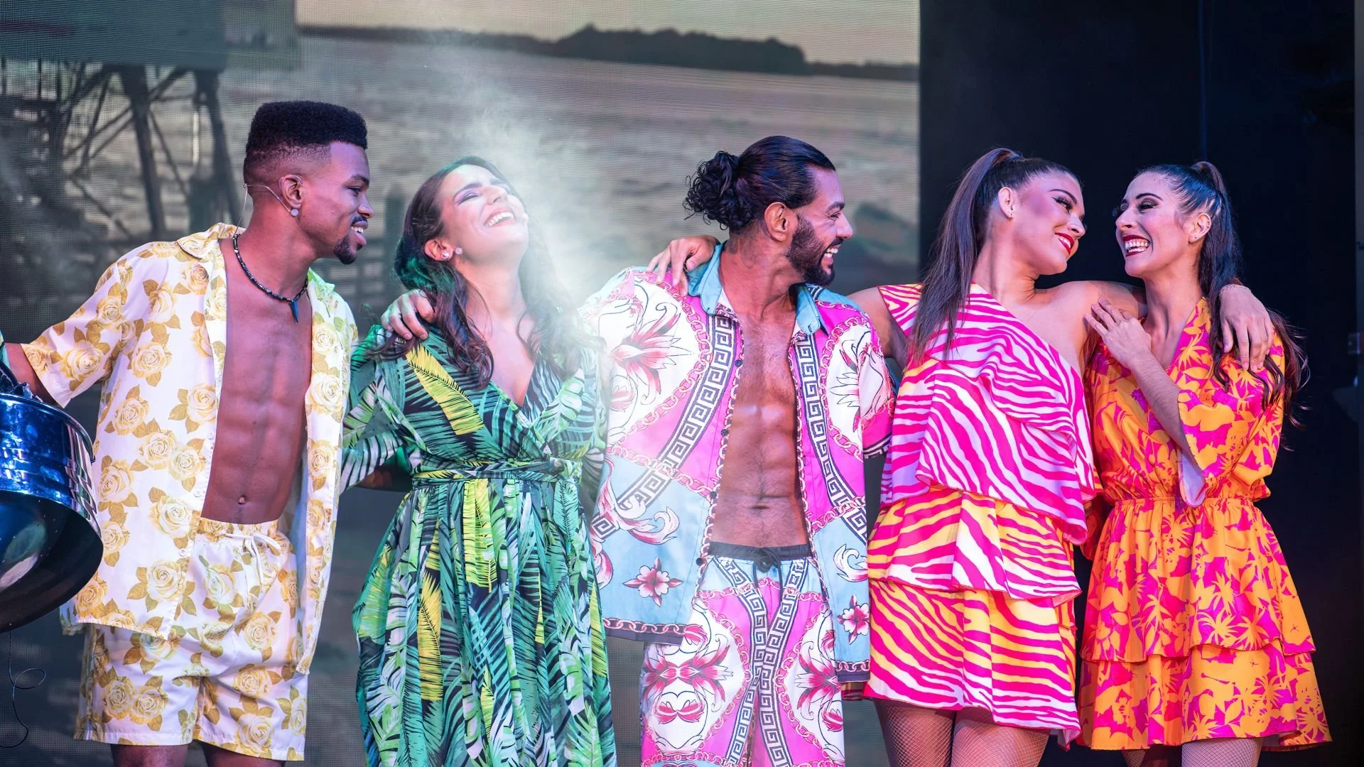 Six people in colorful tropical-themed outfits smiling and standing together on a stage, celebrating.