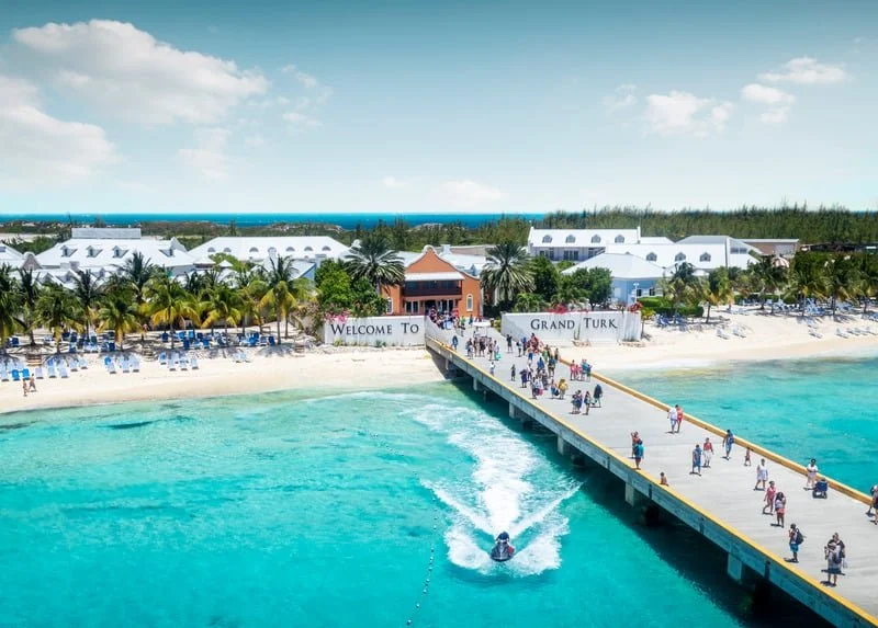 A tropical beach with white sand, palm trees, and lounge chairs. A pier extends into turquoise water with people walking and a jet ski speeding past. Buildings and signs that say 'Welcome To Grand Turk' and 'Grand Turk' are visible in the background.