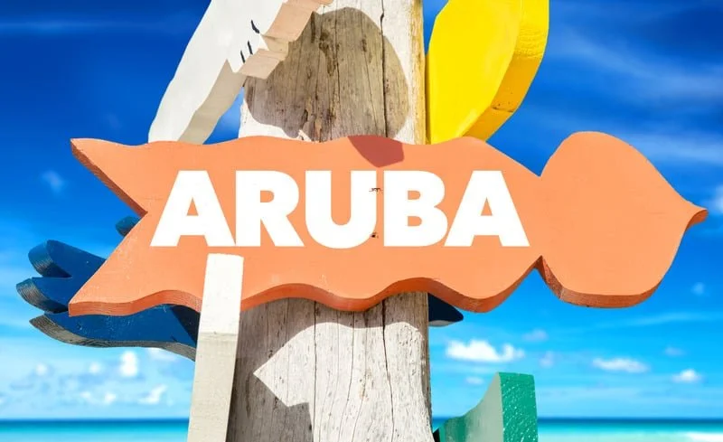 Colorful wooden sign with the word 'Aruba' on a beach with blue sky and ocean in the background.