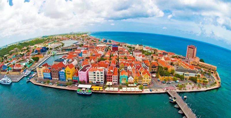 Aerial view of colorful buildings on a harbor island with a bridge connecting it to the mainland, and the ocean in the background, with a partly cloudy sky.