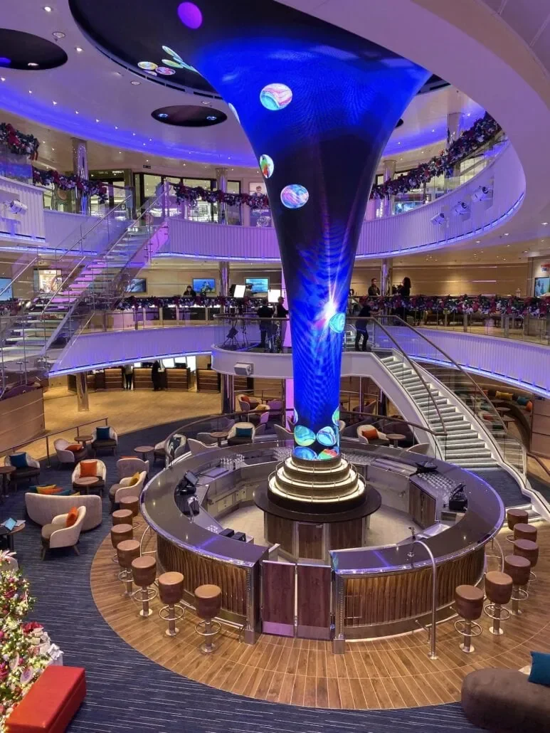 An indoor multi-level lounge or bar area with a large, twisting LED screen feature in the center, surrounded by seating, balustrades, and a staircase. The space is decorated with holiday garlands and has a modern, vibrant atmosphere.