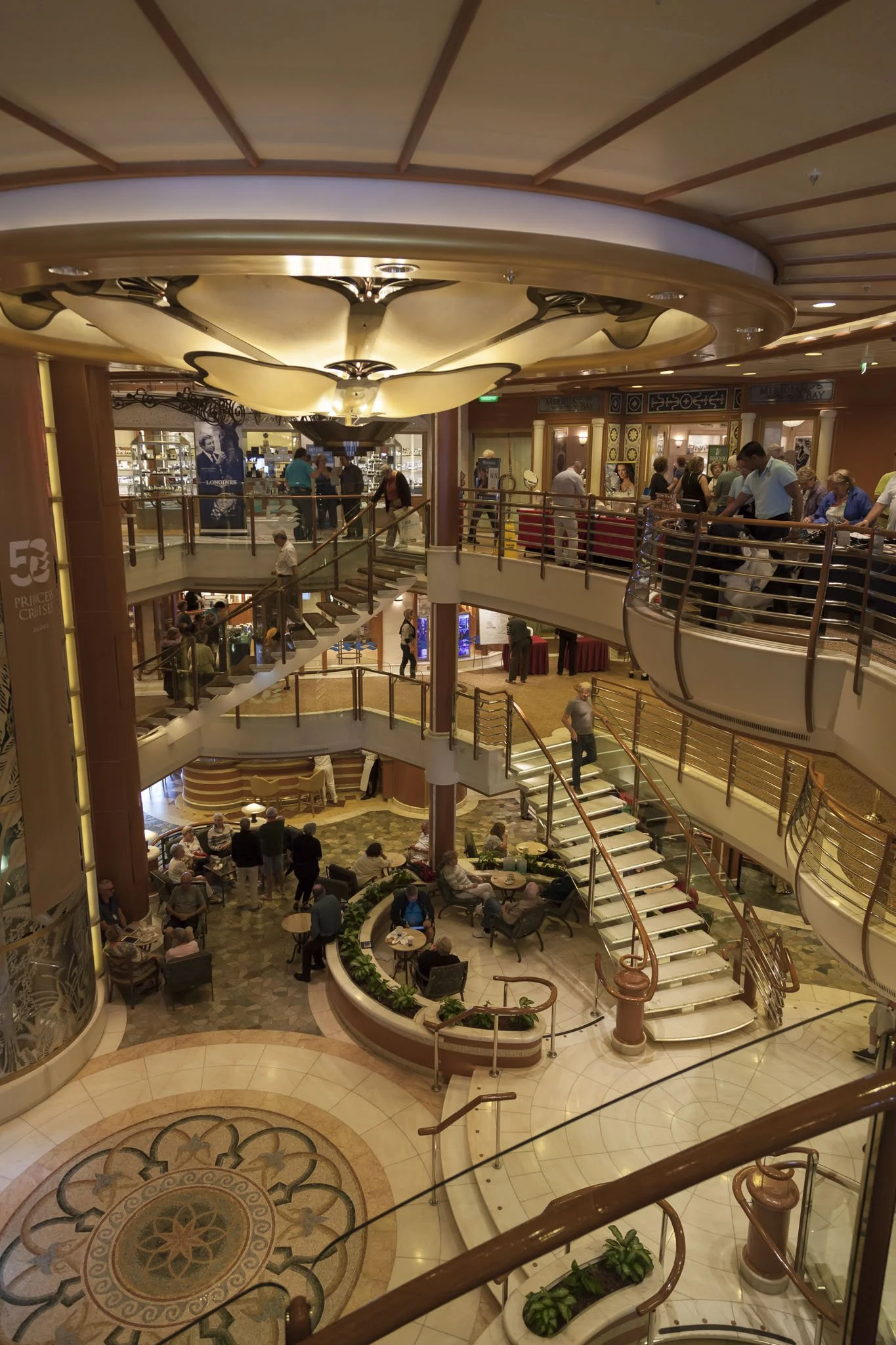 Interior of a multi-level shopping mall with people walking on the upper balcony and stairs, seating area with people, decorative flooring, and a shop at the top level.