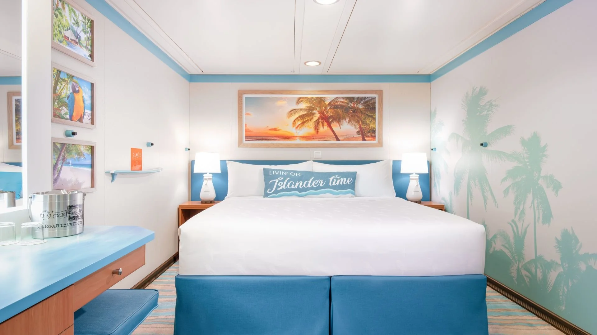 A hotel room with blue and white decor featuring palm tree and island-themed artwork, a bed with white linens and a blue pillow that reads 'LIVIN' ON Islander time,' two white bedside lamps, and a sunset beach scene painting above the headboard.