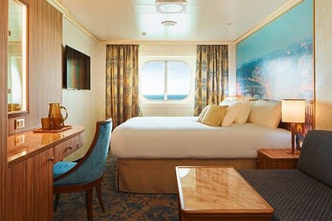 Hotel room with a bed, a window with curtains, a television on the wall, a desk with a chair, and a small sofa with a coffee table.