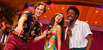 Three young people smiling and laughing on stage, one holding a microphone, in a lively indoor setting with warm lighting and tropical decor.