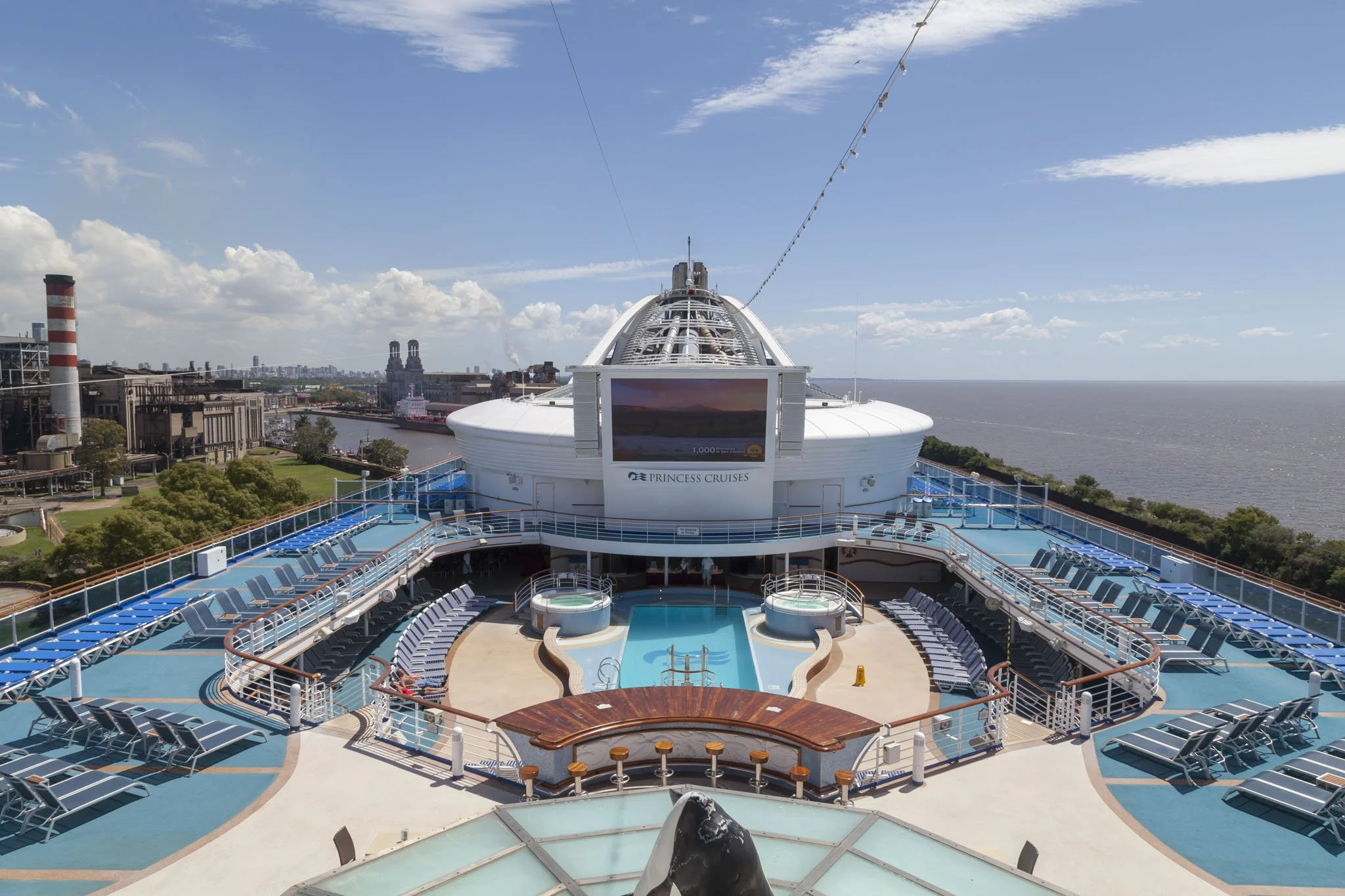 Top deck of a cruise ship with a small pool, lounge chairs, and a large screen, with city skyline and river in the background.